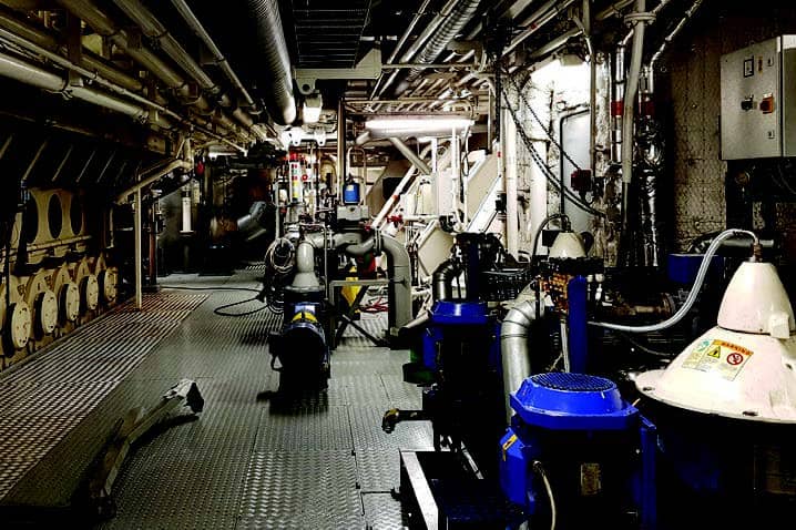 Fotografía de una sala de máquinas de un barco por dentro, con máquinas de distinto tipo y tamaño
                    y tuberías y cables.