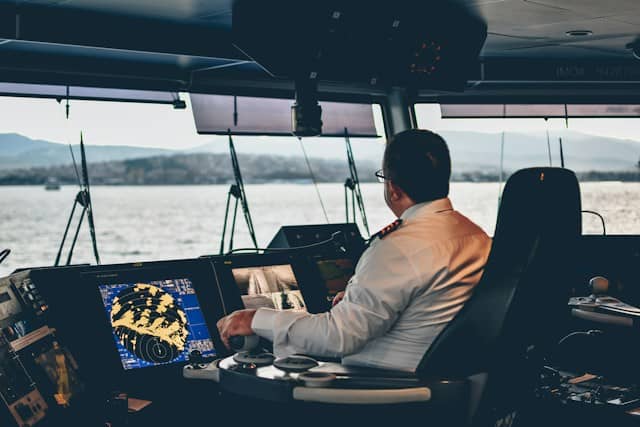 Fotografía de un capitán en la cabina de un barco, manejando con sus controles. Por la ventana del
                    barco se ve el mar y montañas.